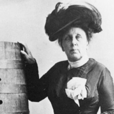 cropped headshot from a black and white photo of a mature looking Annie Taylor in dress and hat standing next to the barrel she went over the falls in