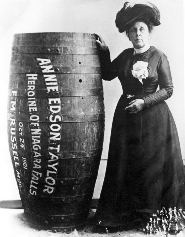 black and white photo of a mature looking Annie Taylor in dress and hat standing next to the barrel she went over the falls in