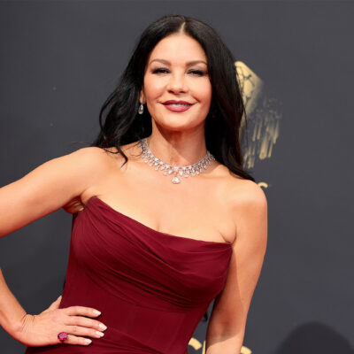 Catherine Zeta-Jones smiles with a hand on her hip in a strapless, dark red gown.