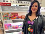 Cece Meadows smiles and stands in front of a makeup display. She is wearing a floral dress with a black leather jacket