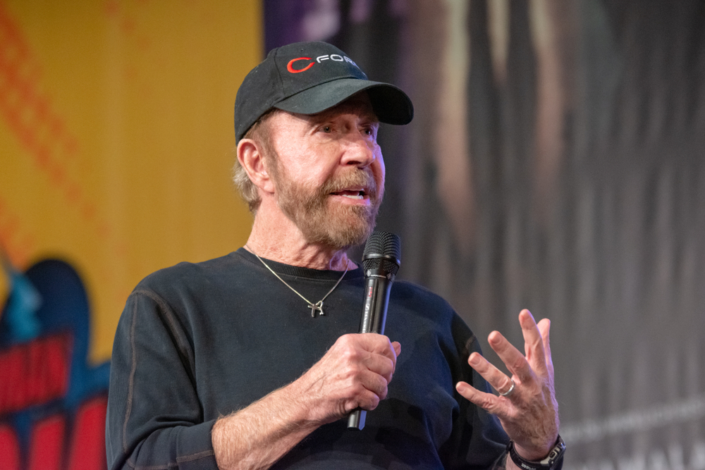 Chuck Norris gives a speech to a crowd wearing a black hat and black shirt. 
