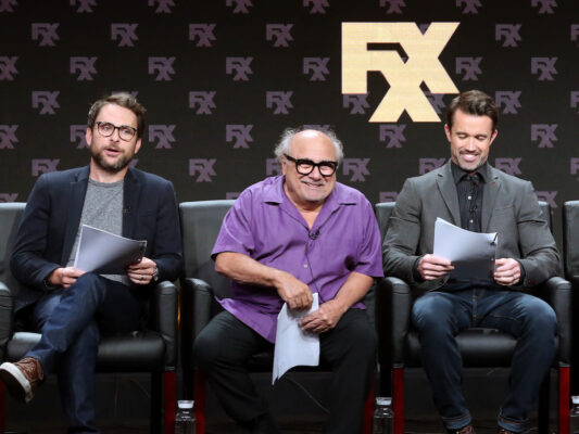 Danny DeVito sitting between Charlie Day and Rob McElhenney on stage during a panel