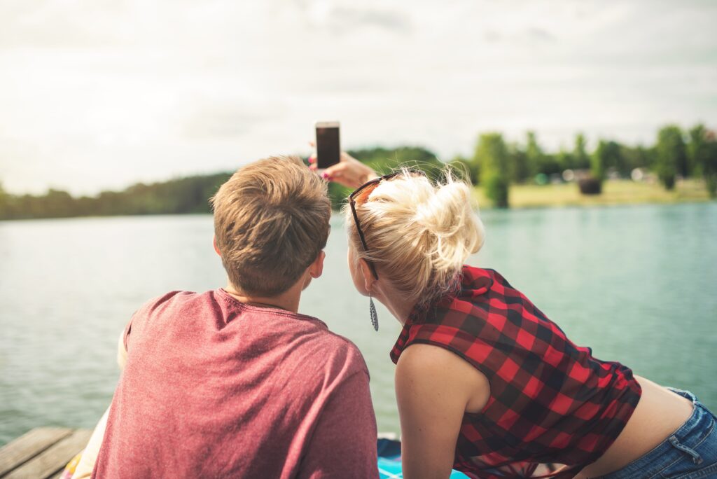 Image of a couple taking a selfie together while sitting on a dock and looking out at a lake.