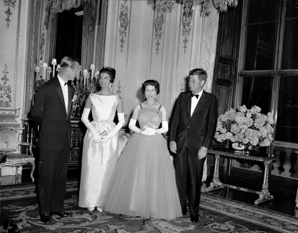(L-R): Prince Philip, Jackie Kennedy, Queen Elizabeth, and John F. Kennedy pose in a black and white photo at Buckingham Palace. All are dressed formally