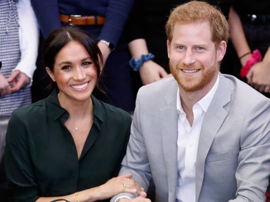 Meghan Markle (L) and Prince Harry sitting and smiling with their arms linked