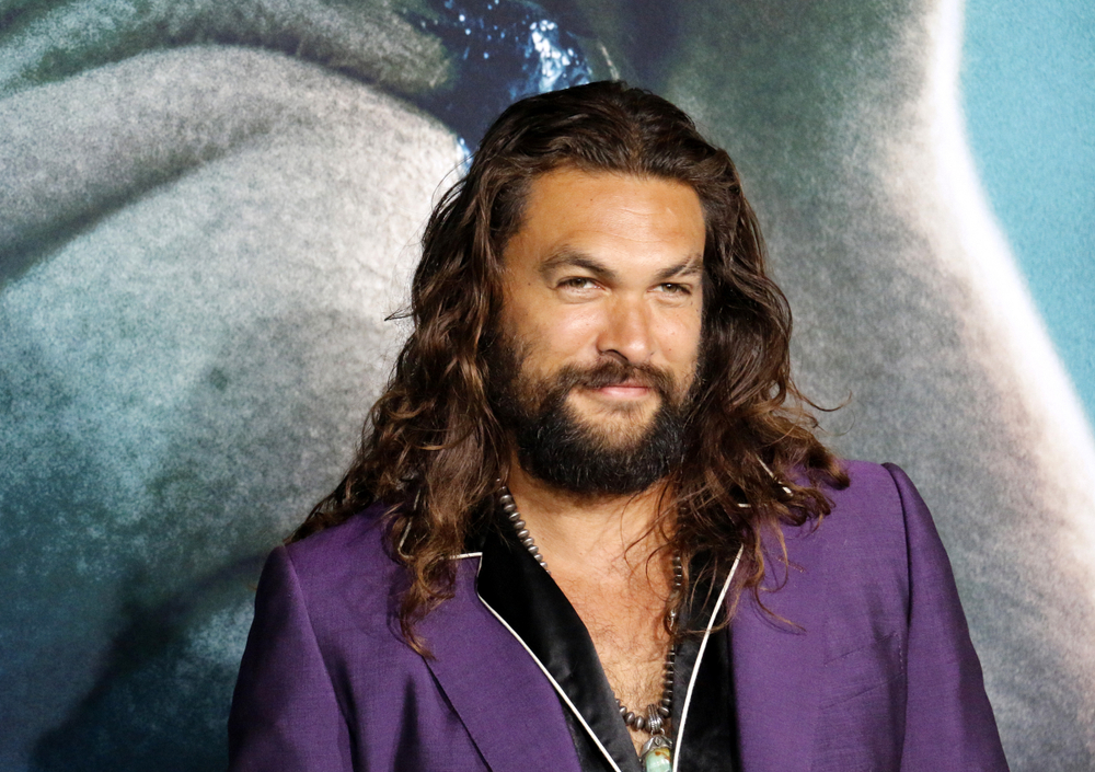 Jason Mamoa poses for the camera, his long hair down, wearing a purple suit and black shirt. 
