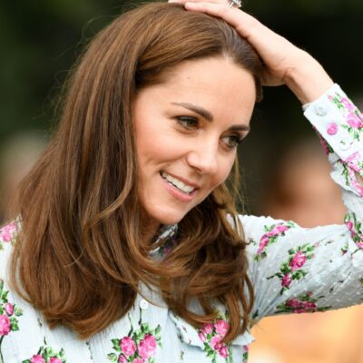 Kate Middleton runs a hand through her hair. She is wearing a white top with pink flowers on it