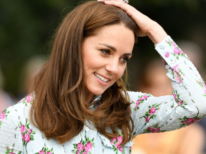 Kate Middleton runs a hand through her hair. She is wearing a white top with pink flowers on it