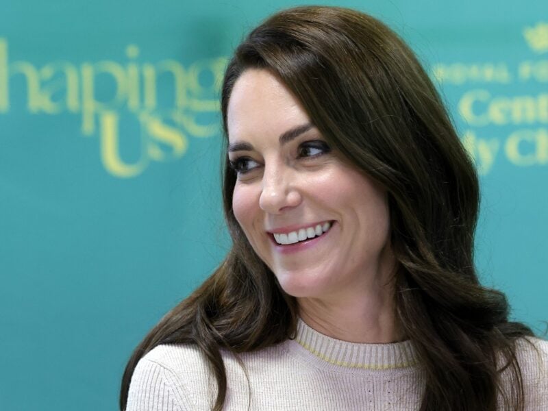 Closeup image of Kate Middleton smiling in beige sweater against teal backdrop