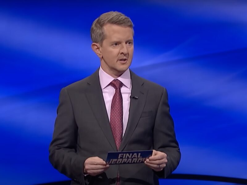 screenshot of Ken Jennings hosting Jeopardy! in a gray suit with his mouth slightly open
