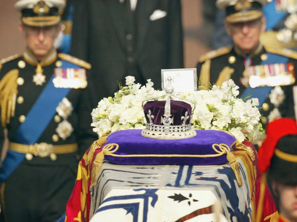 The crown of the queen mother sitting on top of her casket at her funeral with Prince Charles on the left behind the casket and Prince Andrew on the right behind it. 