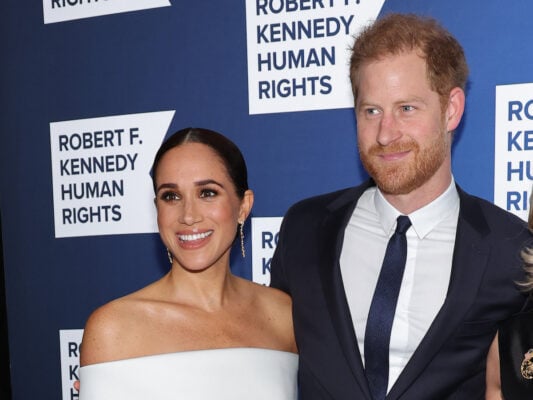Meghan Markle smiling in a white dress with arm around Prince Harry in a black suit