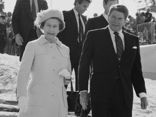 Queen Elizabeth II and Ronald Reagan walking in front of several men in suits, both looking at the camera