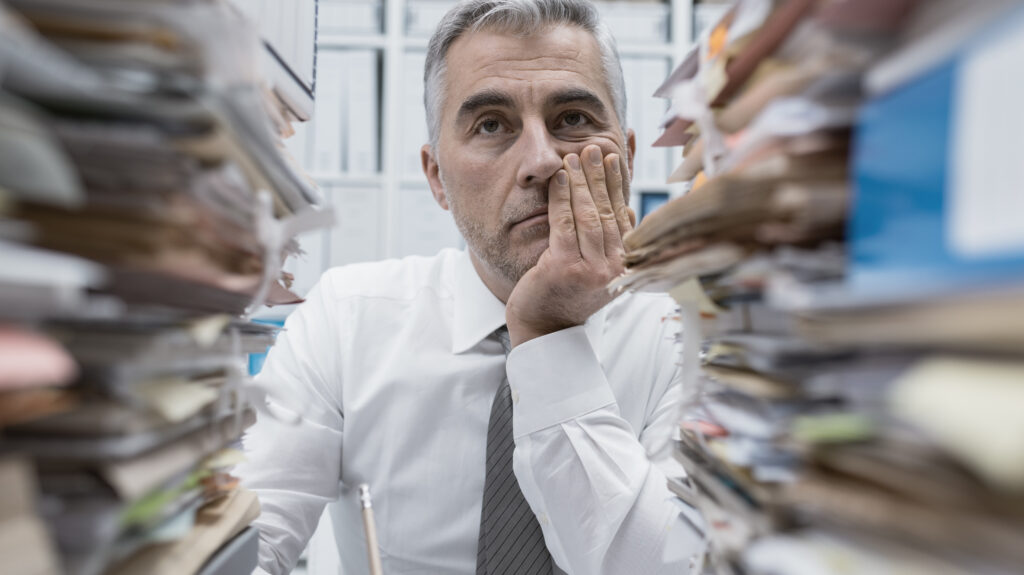 Tired looking man sitting behind large stacks of files and papers. 