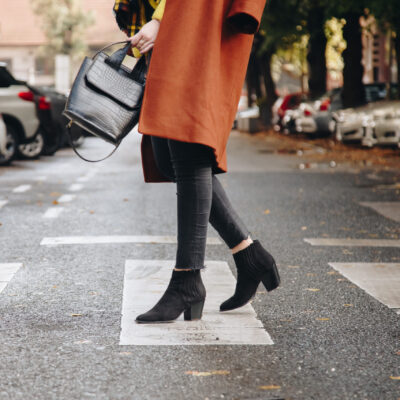 a woman crosses the street wearing black booties and an orange coat