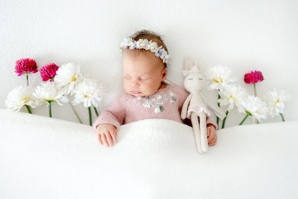 A baby girl sleeps under a white cover and is surrounded by flowers that are tucked in with her