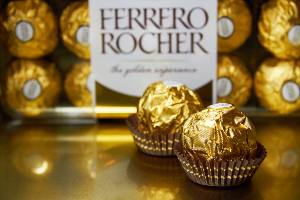 Two Ferrero Rochers classic chocolates in their signature wrappers in front of a box full of chocolates. 