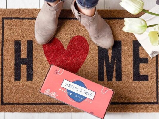 A welcome mat that says "HOME" with the O as a red heart with two boots standing on it and a pink box that says "SinglesSwag" sitting in front of it