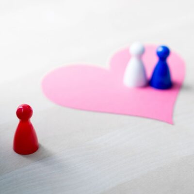 Two board game pawns atop a paper heart on table with a third pawn set apart