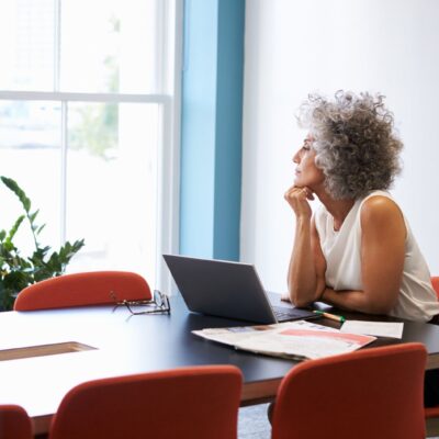 woman with gray curly hair looks out window sitting at desk